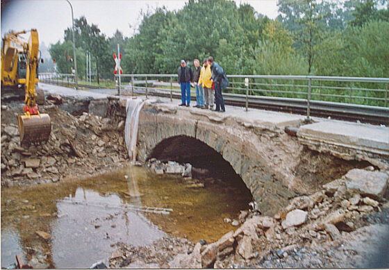 Zerstörte Brücke mit Wasseransammlung und mehreren Personen, die die Situation beobachten.
