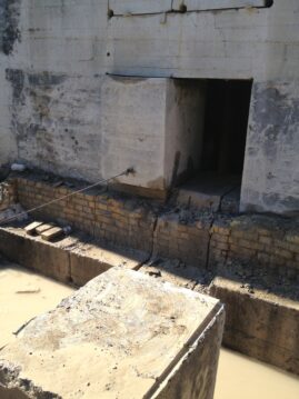 Ein großer Betonblock in einer Baugrube mit feuchtem Boden und einer Wand im Hintergrund.