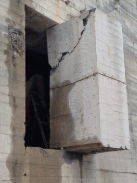Ein großer, rissiger Betonblock, der aus einer Wand herausragt, mit einem Arbeiter auf einer Leiter im Hintergrund.