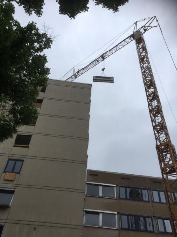 Ein Kran hebt eine Betonplatte an der Seite eines mehrstöckigen Gebäudes in einer städtischen Umgebung.