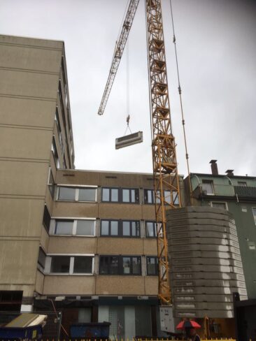 Ein Kran hebt eine Betonplatte auf einer Baustelle in einer städtischen Umgebung.